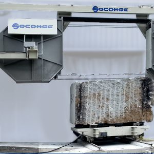 Overhead crane labeled SOCOMEC hoists a large concrete block on a wheeled pallet on a metal framework.