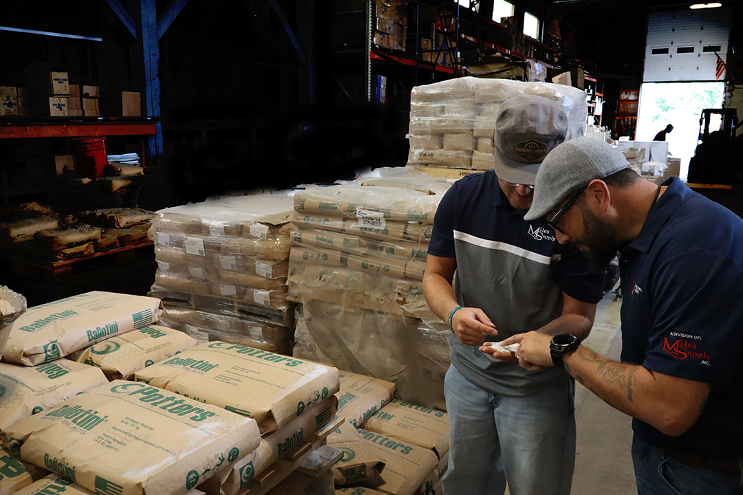 MilesMetal2-LR | Miles Supply Two men in work shirts examine a abrasive grits in their hands inside a warehouse with pallets and bags stacked around them.